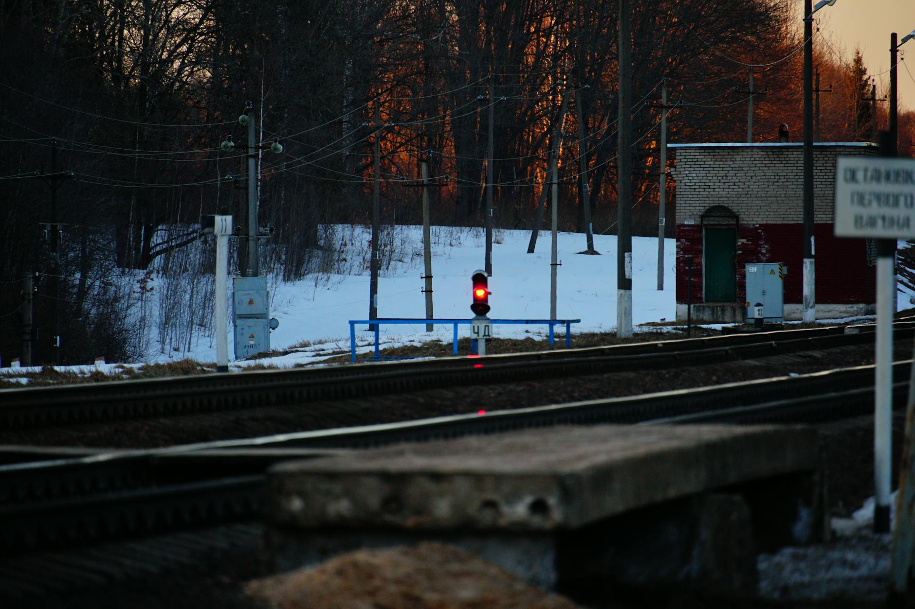 A railway track in winter with a red signal light at sunset, surrounded by snowy trees and infrastructure.