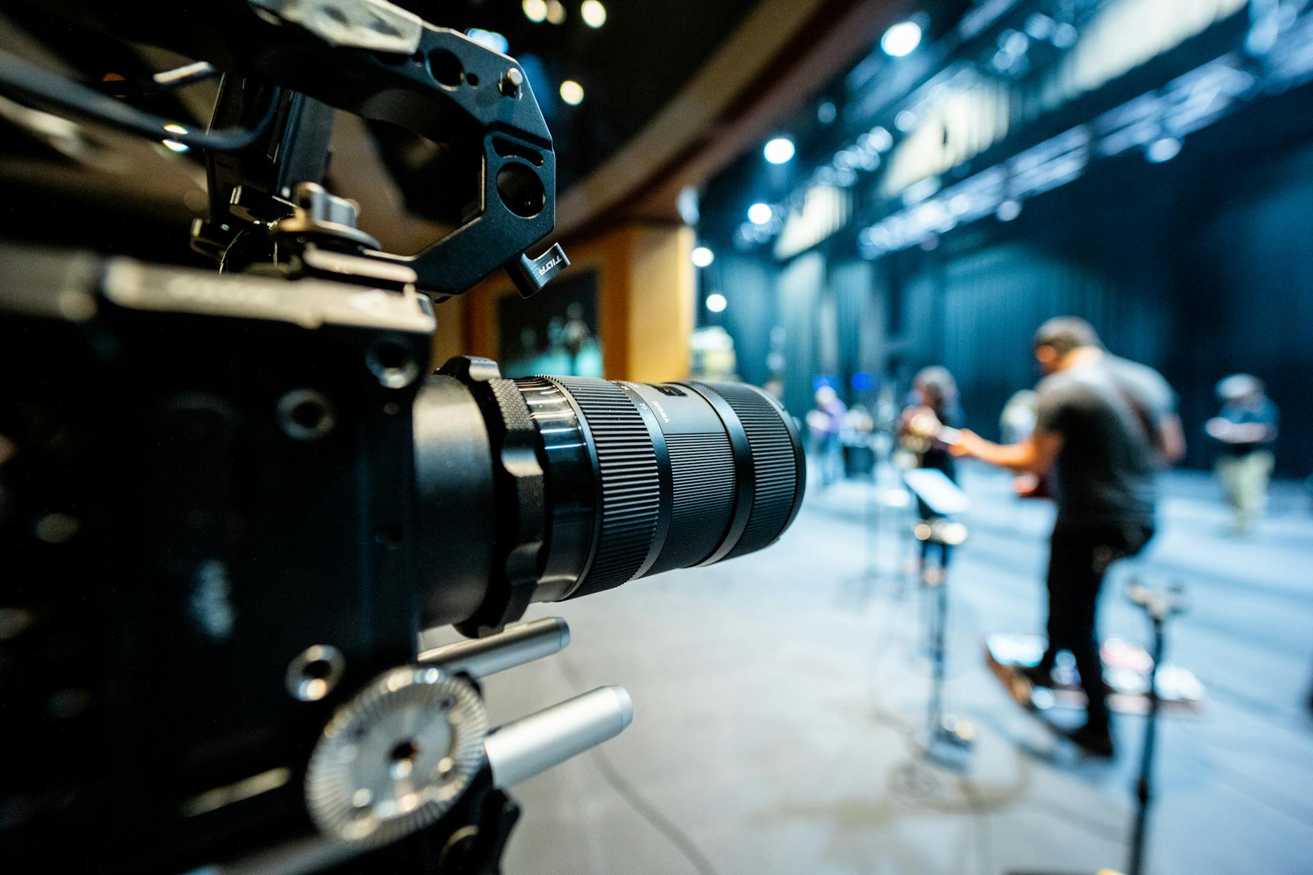 Closeup of professional video camera with lens recording unrecognizable people standing on stage illuminated by lights in studio